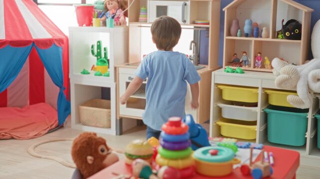 Boy holding a small yellow toy in his right hand while reaching toward a table with stacking rings in a playroom building, surrounded by tent, shelves and storage bins; curiosity.