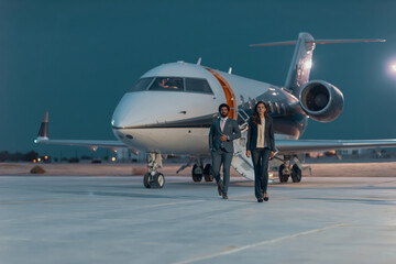 Naklejka premium Confident Middle Eastern business executives in suits walking away from a private jet on an airport tarmac at dusk ideal for luxury business travel and corporate leadership concepts