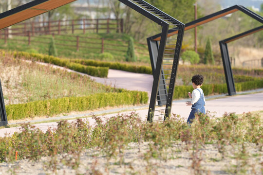 Child explores a modern park with open pathways. Metal arches frame the scene with soft greenery. The child walks curiously, unaware of their surroundings. Bright daylight enhances the tranquil