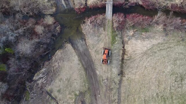 Drone video descending towards an old red forestry skidder near a muddy dirt road crossing a stream on farmland turning yellow in spring.