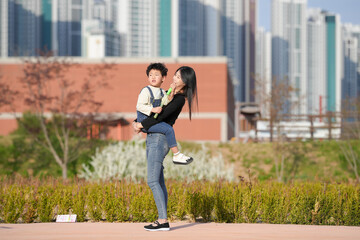 Fototapeta premium Mother hugs child warmly in urban park. Sunlight glows on their joyful faces. Buildings rise softly behind greenery. Perfect moment of love, play, and peace. Great for family, lifestyle