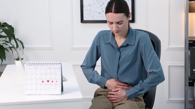 Woman suffering from menstrual pain at white desk with calendar with noted period days. Camera moving forward