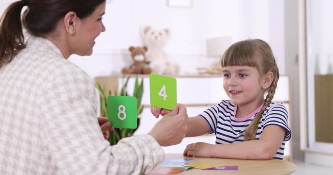 Speech therapist teaching girl numbers with colorful cards at wooden table indoors