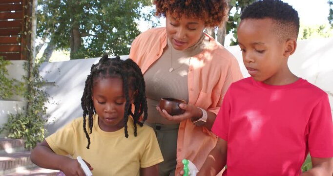 African American woman offering bowl guiding kids planting seeds using sprayers at backyard table