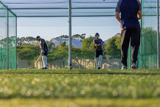 Male teammates practicing cricket in net cage on turf, holding bats, wearing pads and helmet