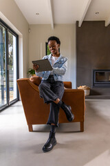 Obraz premium African American woman 20s sitting on leather armchair at home holding tablet, wearing black boots