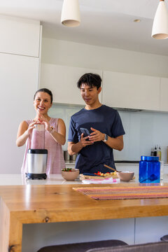 Couple using countertop blender while holding smartphone and preparing fruit on kitchen island