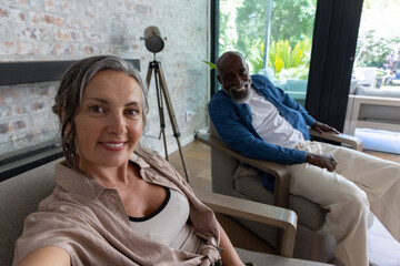 Obraz premium Couple sitting in lounge smiling while woman taking selfie with phone near glass doors, copy space