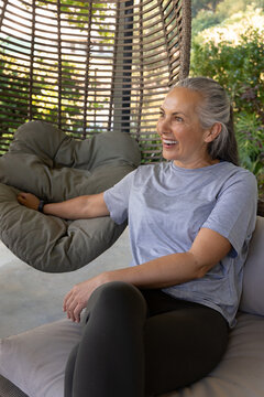 Senior woman sitting in rattan hanging chair on patio, resting arm on cushion, checking wristband
