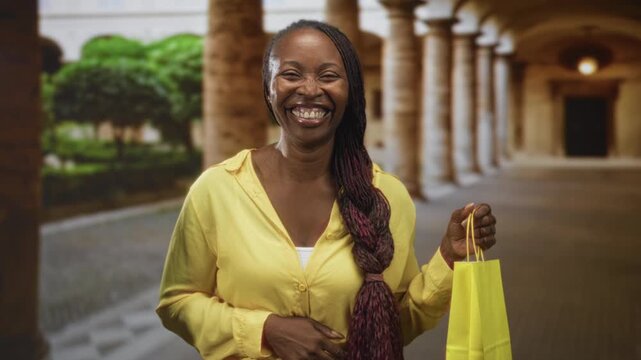 Young african american woman smiling broadly, holding a yellow shopping bag in a building colonnade with braided hair visible; joy.