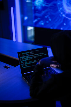 Mysterious hacker in black hoodie stealing confidential data on laptop. Cyber criminal coding dangerous malware virus to attack network system in dark blue room.