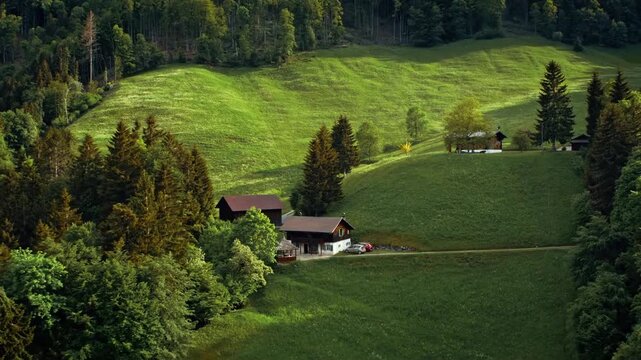 Aerial landscape view of a secluded traditional farmhouse nestled in the lush green rolling hills of the alpine countryside during a serene summer afternoon