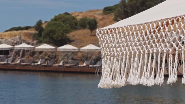 Cinematic 4K shot of a white macrame fringe canopy in foreground with a luxury beach club, wooden deck, sunbeds, umbrellas and rocky hillside in the background. Aegean sea.