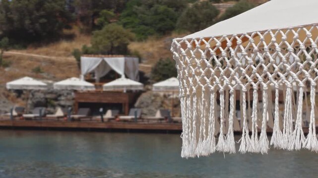 Cinematic 4K shot of a white macrame fringe canopy in foreground with a luxury beach club, wooden deck, sunbeds, umbrellas and rocky hillside in the background. Aegean sea.