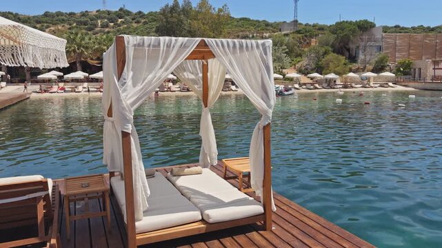 Slow pull-back wide shot of white canopy daybed on wooden pier over turquoise Aegean bay, luxury beach club with sun loungers in background,