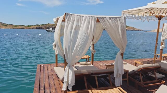 Slow pull-back wide shot of white canopy daybed on wooden pier over turquoise Aegean bay, luxury beach club with sun loungers in background,