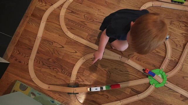 Young boy with red hair kneels on wooden flooring, actively arranging toy trains on a wooden track layout, demonstrating engagement and creativity in a warm indoor environment filled with toys