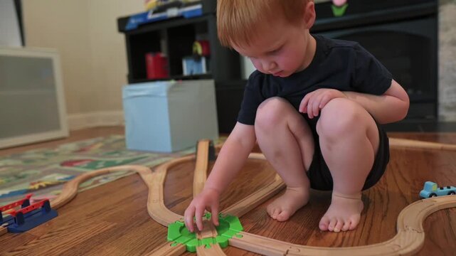 Young boy with red hair crouches on a wooden floor, carefully placing wooden train tracks with green connectors, while colorful toy vehicles are scattered around in a bright indoor environment