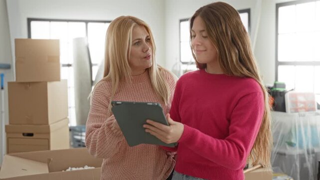 Two women mother and daughter hold a tablet and laugh while one raises her hand beside stacked moving boxes in a bright building interior; family joy.
