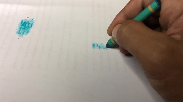 A hand holding a blue-green crayon to color a small square pattern on white paper, leaving behind bright, textured wax particles.
