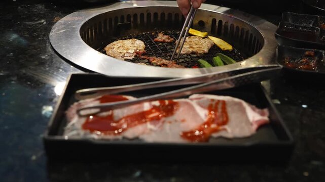Raw sliced pork with red sauce in a black tray with tongs. In the background, meat and vegetables grill on a circular inset metal grate. Authentic Asian barbecue restaurant dining scene.