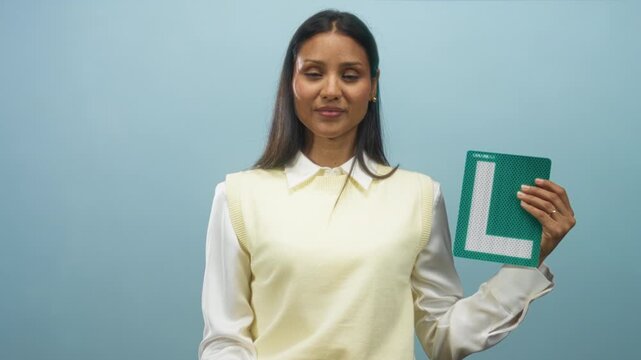 Woman young hispanic brunette wearing pale yellow vest and white blouse holding green l plate and points fist forward in studio; confidence success.