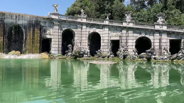 Smooth lateral tracking shot of the historic baroque fountain and tiered stone arcades at Reggia di Caserta, capturing water features, statuary, and reflecting pool