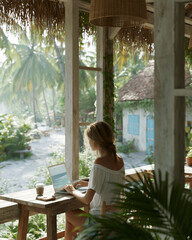 Obraz premium Woman works quietly by a rustic porch. Laptop sits on a weathered wooden table. Dappled sunlight filters through thatched roof. Palm trees sway softly in the background breeze