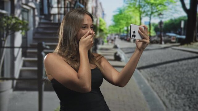 Young woman covers mouth with hand while holding smartphone for selfie on a street near stoop stairs and sidewalk; surprise personal moment.