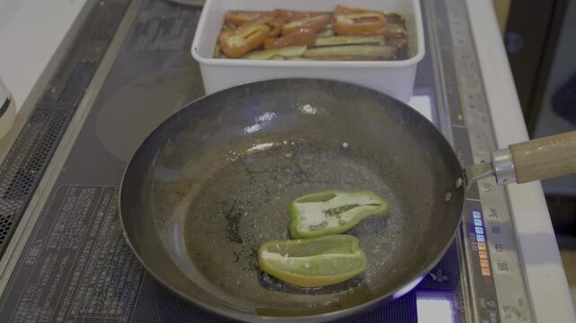 煮浸しで緑色のピーマンを鉄フライパンに入れ、IHクッキングヒーターの中火で焼き上げるシーン