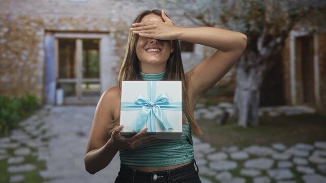 Woman holding white gift box tied with blue ribbon, right hand covering eyes, smiling, standing on cobbled path in front of stone building; surprise celebration.