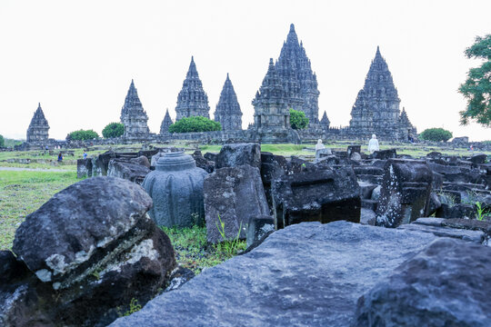 Ancient temple ruins stretch across green grassy terrain. Weathered stone blocks lie scattered in foreground. Five pointed spires rise against pale sky. Lush tree provides contrast to historic stones