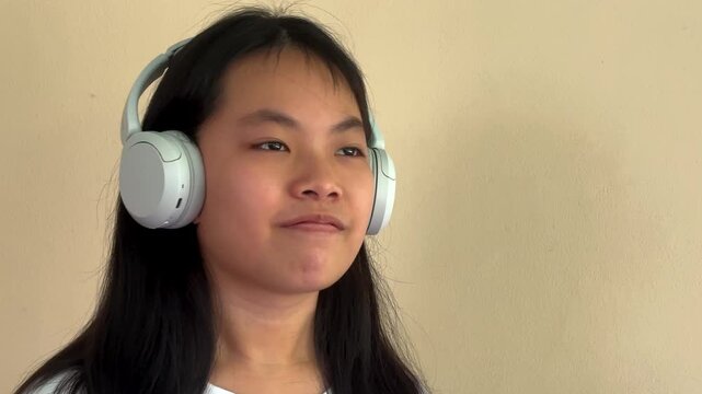 A static, medium close-up shot capturing a preteen Asian girl completely immersed in her favorite music. She wears large white over-ear wireless headphones, displaying a subtle, feel-good expression
