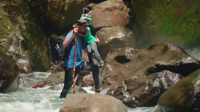 Son with father hiking wild nature as they navigate rushing river waters between mossy boulders. Dad uses walking stick while guiding child across slippery rocks in lush green canyon with waterfall.