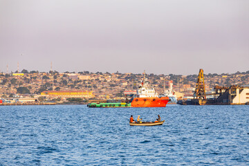Fototapeta premium Local fishermen in small wooden boat on foreground with offshore supply vessel and industrial coastal city on background