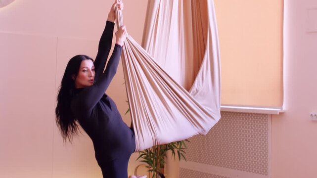 Female training. Muscle strength. Athlete descending from hammock unrolling safety fabric support doing fly yoga side split pose in studio.