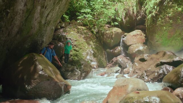 Father and son carefully wade through rushing turquoise waters of a tropical river canyon, using walking sticks for balance among massive wet boulders beneath overhanging rock walls and ferns.