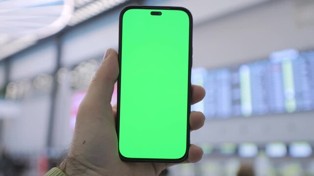 Hand holds phone with green screen at airport. Background shows blurred flight info boards. Person appears focused, checking travel details. Ideal for travel, tech, or communication concepts