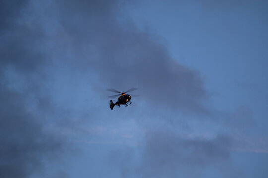 Police helicopter in flight viewed from below against an overcast blue-grey sky with searchlight illuminated
