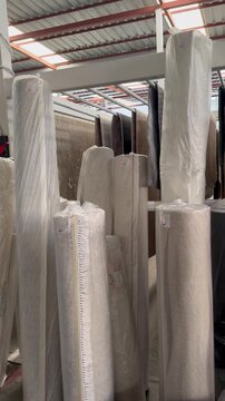 Tall rolled mattresses and wrapped bedding materials are arranged in neat rows inside a covered retail storage space, with carpets and other home furnishings