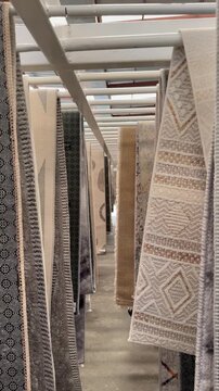 Rows of decorative textile samples hang from overhead racks in a narrow indoor aisle, creating a symmetrical view of patterned rugs in soft warehouse lighting