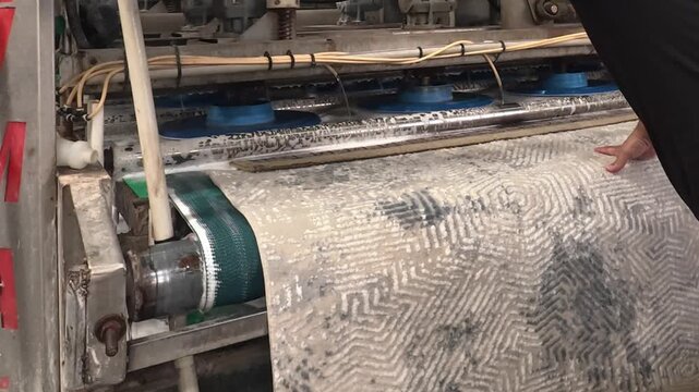 A patterned carpet moves through industrial rollers while a worker applies liquid in a bright factory setting, showing textile processing, maintenance