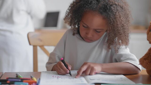 Cute little preschool or primary student girl doing homework in notebook at home. Portrait of pretty african american child in kitchen, mother talking by phone, walking in background, modern family