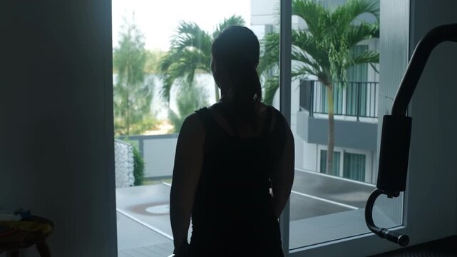 Dark silhouette of a young woman performing dumbbell lateral raises in a gym with bright natural light coming through large windows behind her, slow motion. Fitness concept