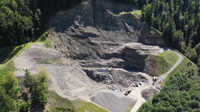 Aerial view of an active quarry surrounded by forest. Exposed rock faces, excavation terraces, gravel stockpiles, access roads, and heavy machinery highlight industrial mineral extraction operations.