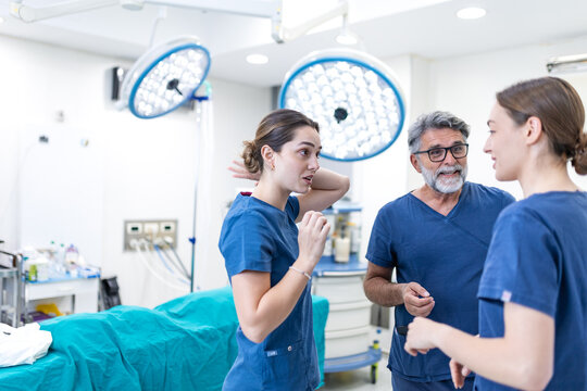 Three medical professionals in blue scrubs standing inside a modern operating room without masks and caps, engaging in a casual, smiling conversation and discussion after a successful surgery.