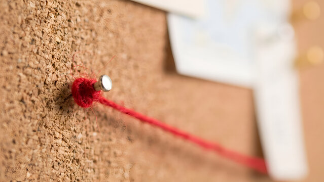 A close-up of a red push pin holding a red string on a brown cork bulletin board, with blurred papers in the background, representing connection