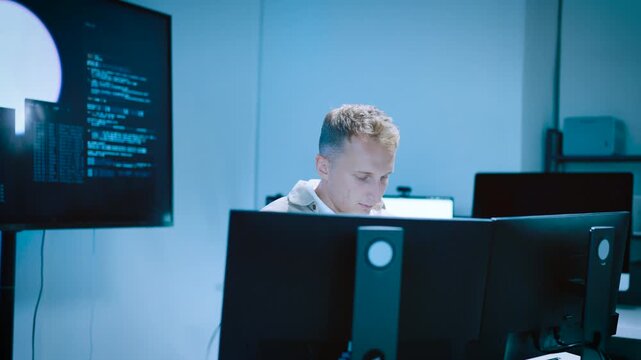 Focused male developer analyzing code on multiple desktop monitors in a modern server control room with blue ambient lighting
