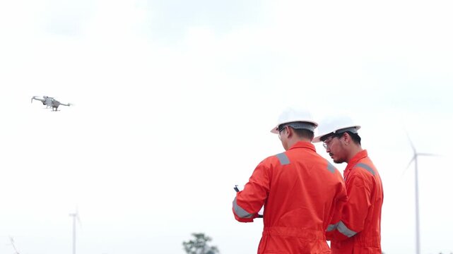 Focused Asian utility operators tracking automated inspection drone inside turbine power field. Two male safety engineers reviewing clean alternative electricity plant solution.