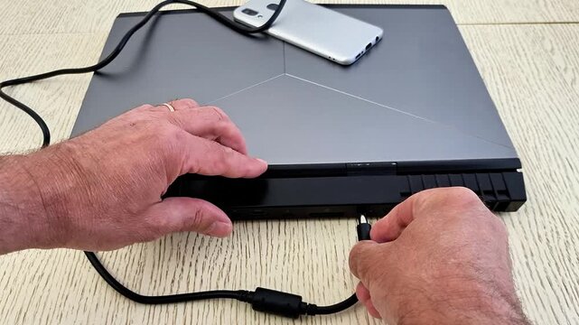 Hands connect cable to back of metallic gray laptop with angular design, smartphone resting on top, showcasing modern tech setup for charging or data transfer on textured table
